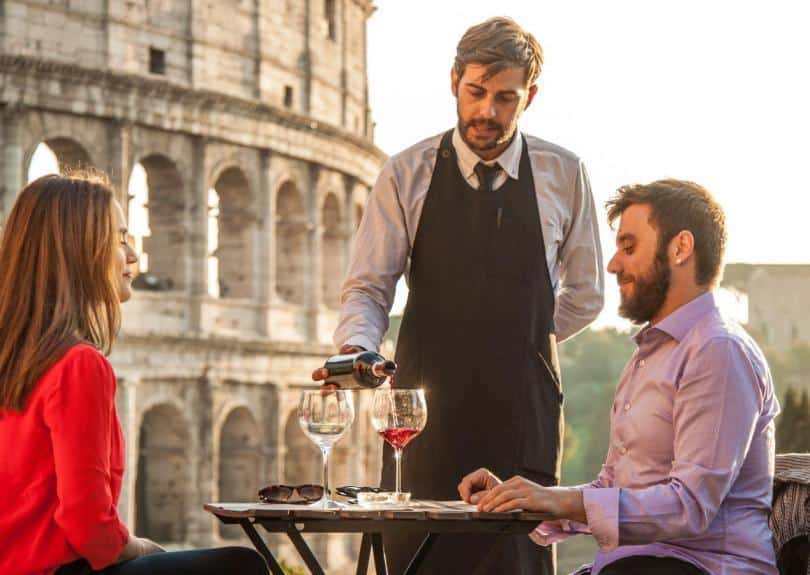 Elegant waiter serving red wine to a young happy couple in bar restaurant in front of colosseum in rome at sunset
